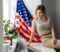 Happy woman employee sitting wrapped in USA flag, shouting for joy in office workplace, celebrating labor day or US Independence day. High quality photo