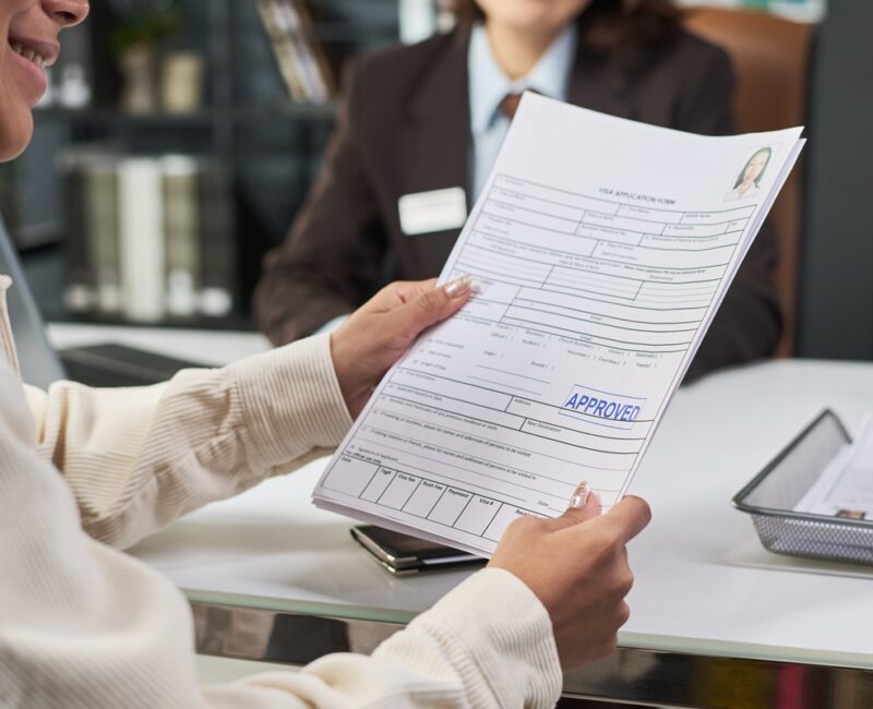 Young adult Latin woman holding approved visa application form while sitting across from middle aged Caucasian woman at visa center desk, both engaging in document review process