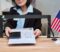 Caucasian middle aged woman handing approved visa application form and passport through service window at visa center, American flag standing on counter, partial face visible
