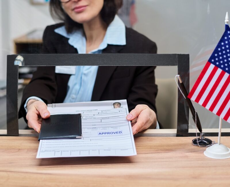 Caucasian middle aged woman handing approved visa application form and passport through service window at visa center, American flag standing on counter, partial face visible